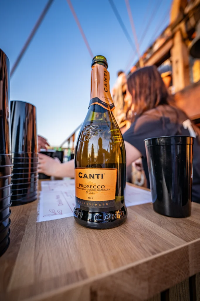 Canti Prosecco bottle on a wooden bar table at an outdoor event, with people and plastic cups in the background