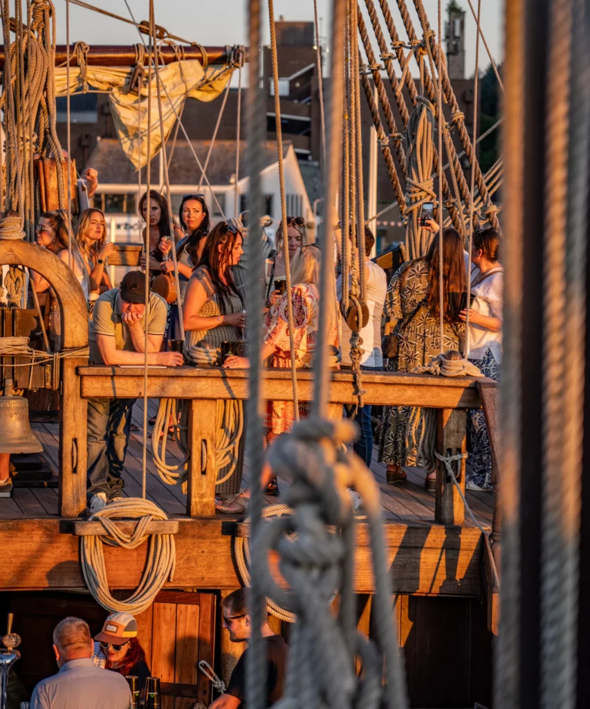 Guests socialising on the deck of a historic wooden ship in bright daylight, with rigging and ropes in the foreground.