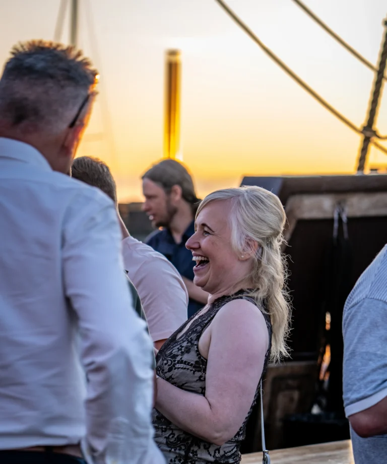 Guests laughing and chatting on the deck of a tall ship at sunset
