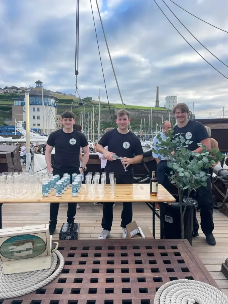 Three smiling bartenders on the deck of a historic ship serving drinks at an outdoor harbour event