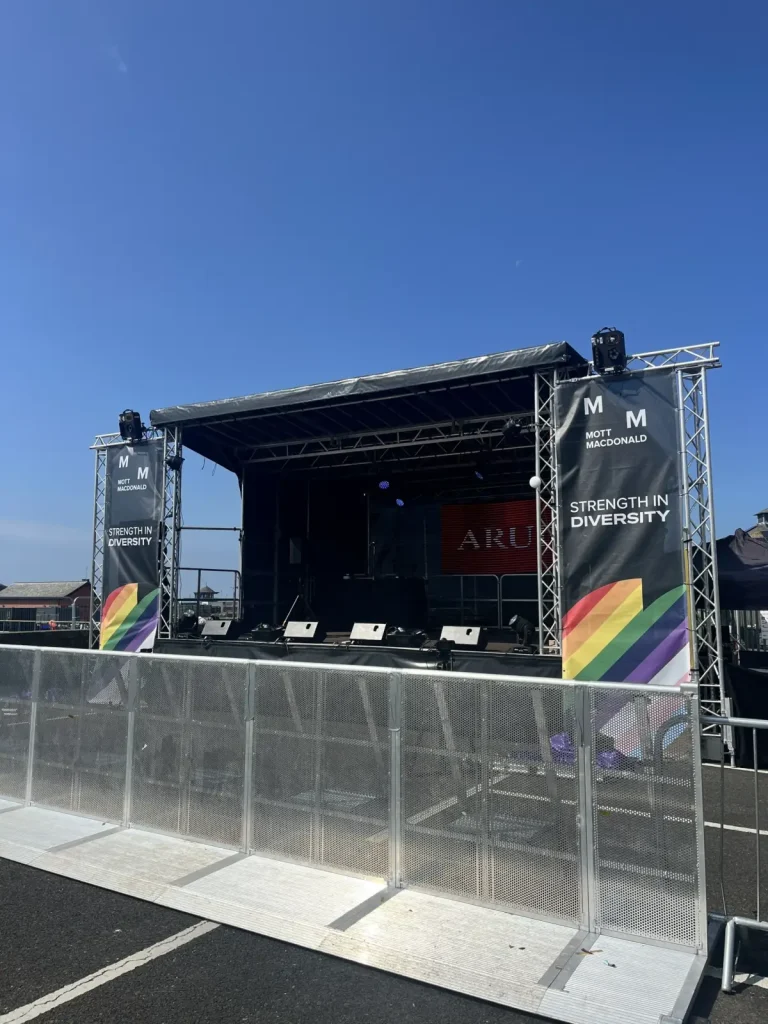 Outdoor stage with lighting rig and rainbow Strength in Diversity banners behind crowd barriers