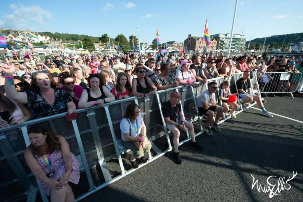 Crowd behind barriers waving rainbow flags at an outdoor Pride event stage