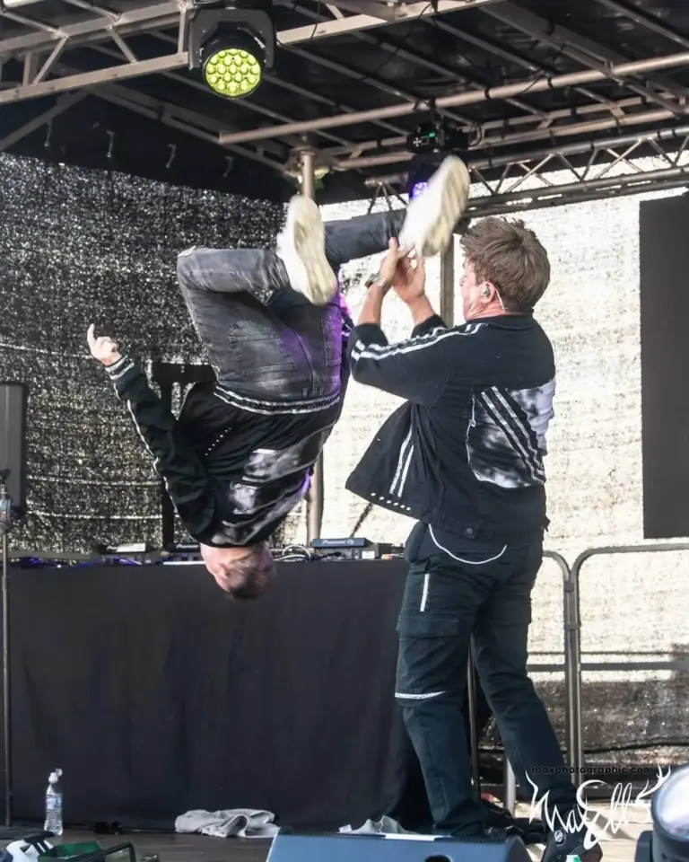 A performer doing an upside-down handstand stunt with a DJ on an outdoor stage