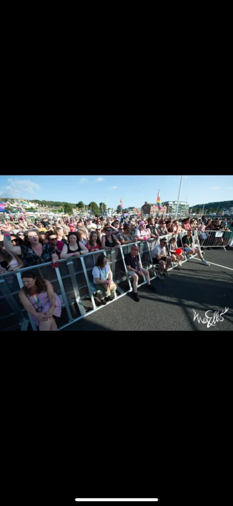 Large crowd gathered behind safety barriers at an outdoor Pride event, watching the stage on a sunny day.