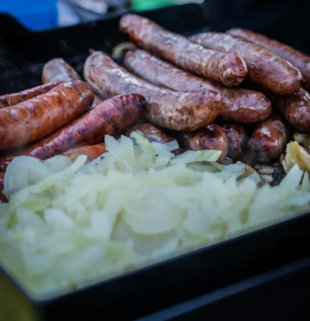 Grilled sausages cooking on a hotplate with a pile of sliced onions.